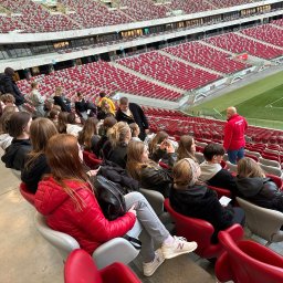 Biuro Turystyczne Skaut Tour sp. z o. o. - Wycieczka szkolna na stadionie: grupa młodzieży siedzi na trybunach, słuchając przewodnika w czerwonej kurtce. W tle widoczne czerwone krzesełka i boisko.