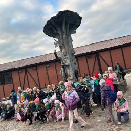 Biuro Turystyczne Skaut Tour sp. z o. o. - Grupa dzieci w zielonych chustach pozuje przed fontanną w kształcie drzewa z tabliczkami kierunkowymi, ceglany budynek w tle. Pochmurne niebo.
