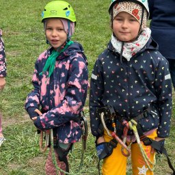 Biuro Turystyczne Skaut Tour sp. z o. o. - Dwie dziewczynki w kaskach i uprzężach wspinaczkowych stoją na trawie. Jedna ma zielony kask, druga biały. Ubrania kolorowe, w tle zielona trawa i fragmenty innych osób.