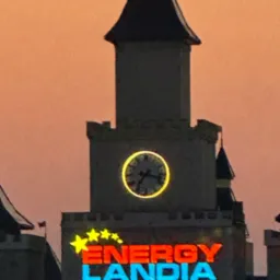 Wieża z zegarem i neonowym napisem Energylandia na tle pomarańczowego nieba o zmierzchu. Widoczne detale architektoniczne zamku.