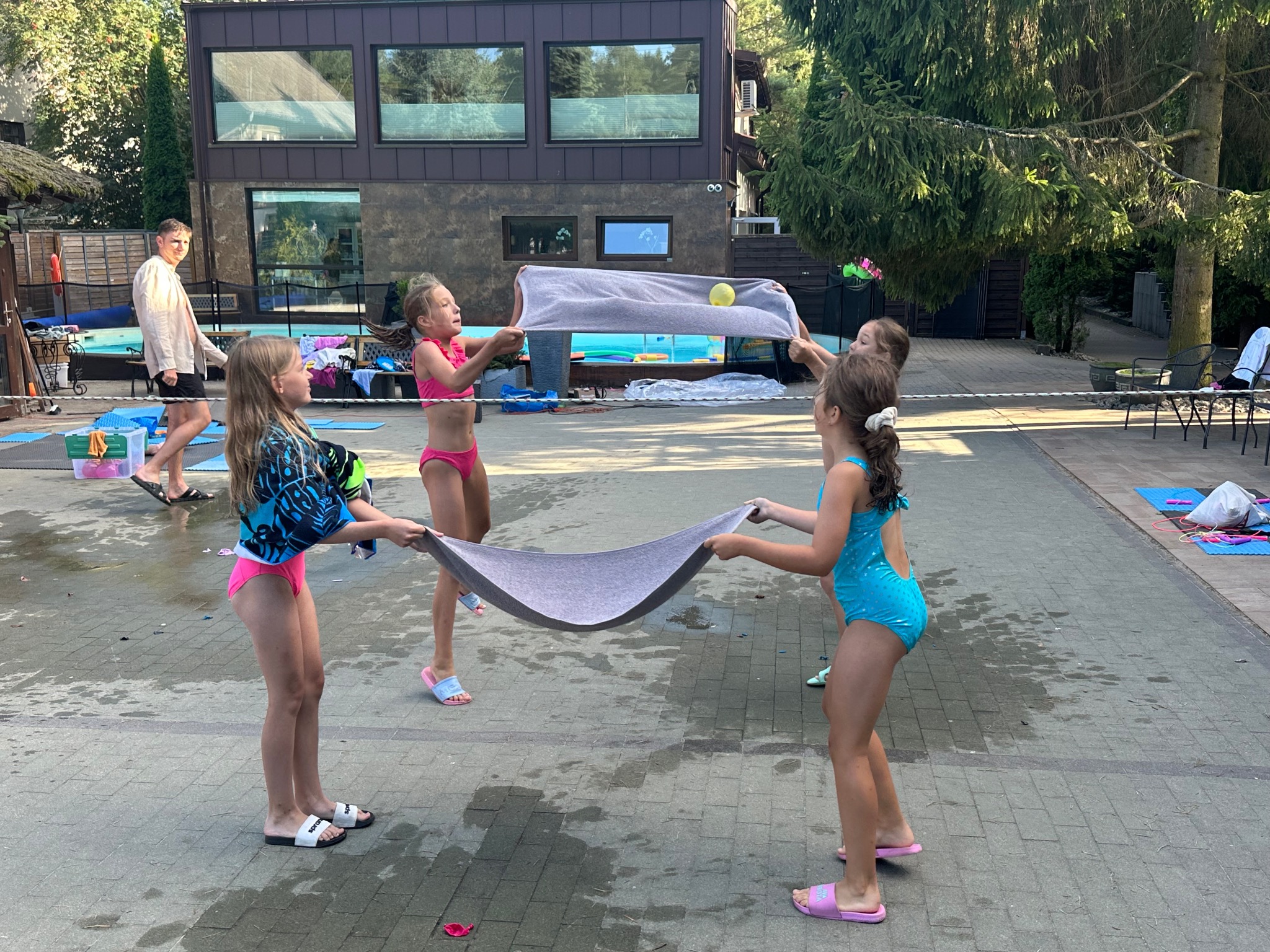 Grupka dzieci w strojach kąpielowych bawi się na zewnątrz, trzymając napięty ręcznik z piłką. W tle basen i budynek z dużymi oknami. Letnia aktywność na świeżym powietrzu.