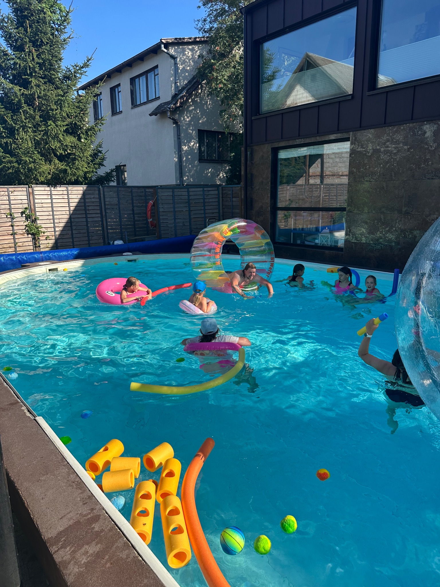 Radosna grupa dzieci bawi się w basenie z kolorowymi zabawkami, dmuchanymi kołami i tunelem. W tle budynki mieszkalne w słoneczny dzień.