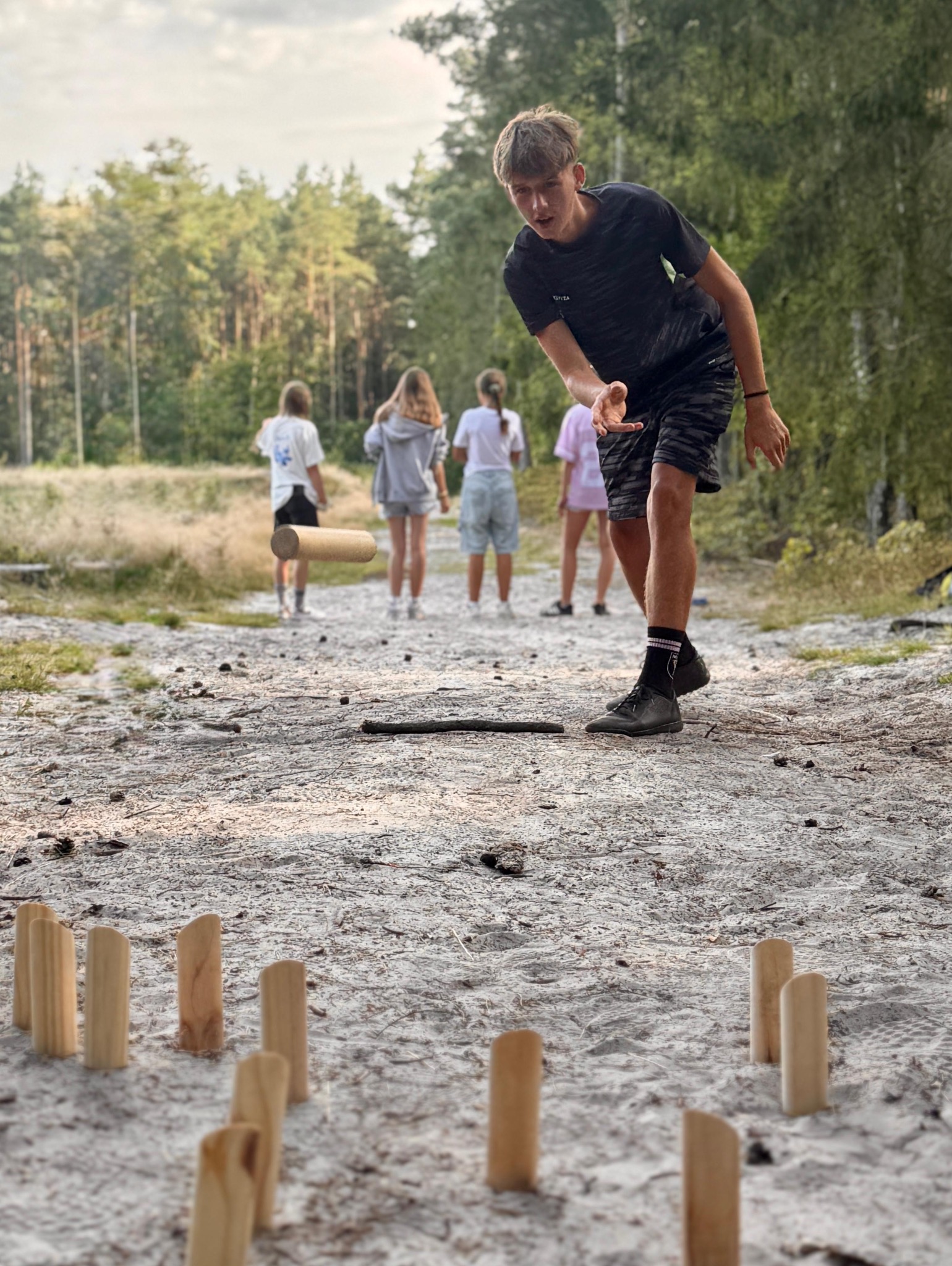 Gra Kubb w plenerze w Łodzi. Młodzieniec rzuca drewnianym kołkiem w ustawione pionowo klocki, w tle grupa osób i las. Aktywność na świeżym powietrzu.