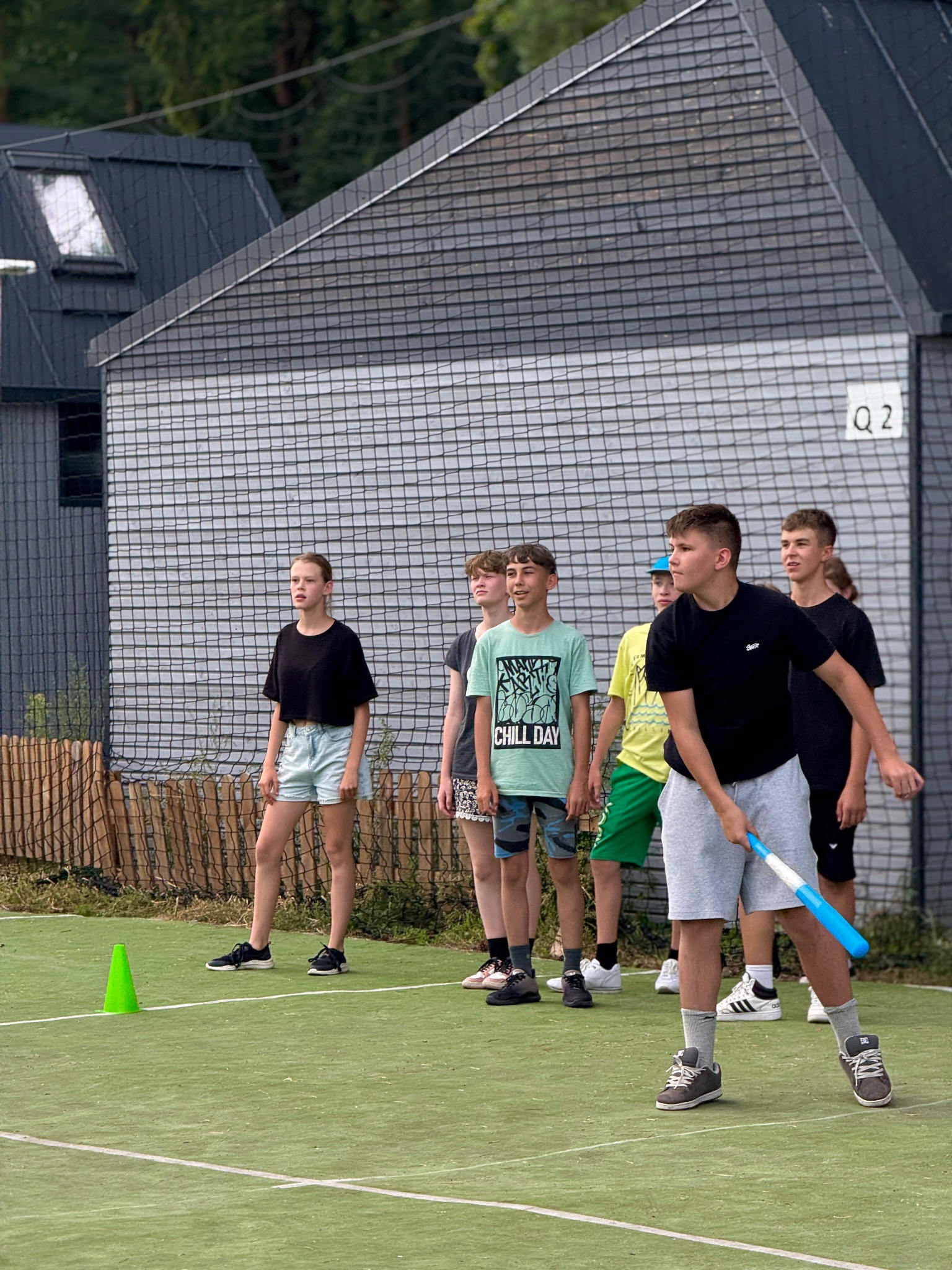 Grupa młodzieży na boisku, jeden z nich trzyma kij do gry. W tle budynek z siatką ochronną. Koncept sportowy, rekreacja na świeżym powietrzu.