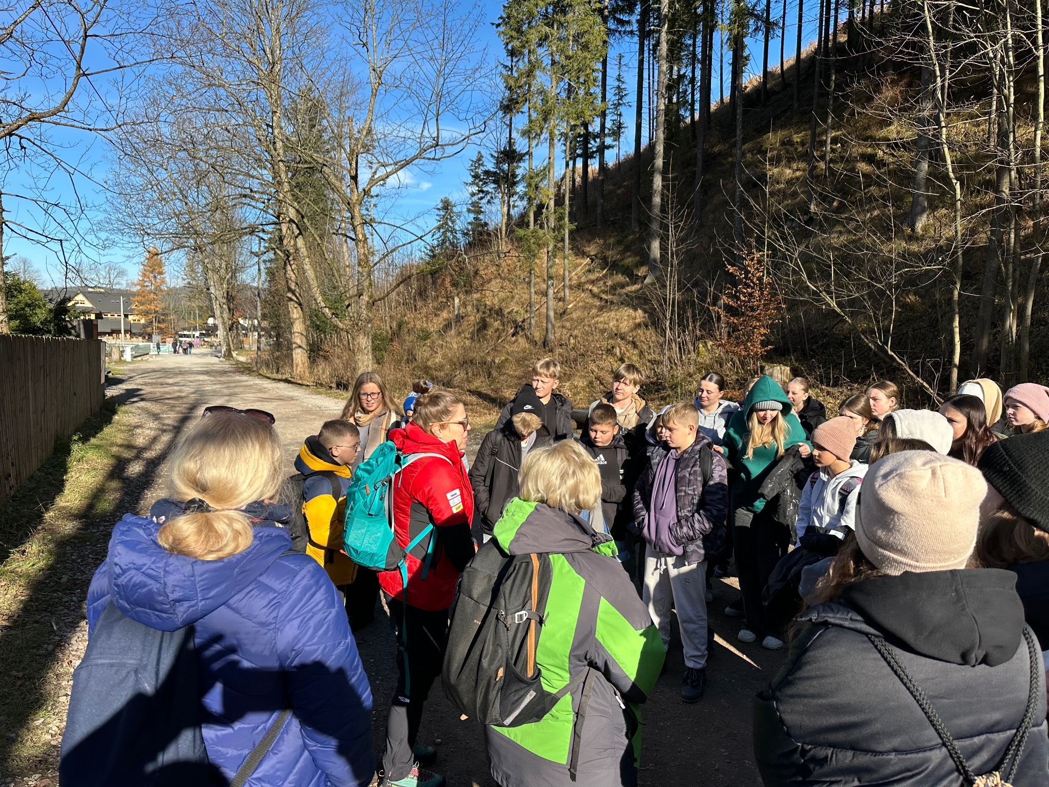 Grupa młodzieży z plecakami, prawdopodobnie podczas wycieczki edukacyjnej w lesie, w słoneczny dzień. W tle widoczna ścieżka i drzewa.
