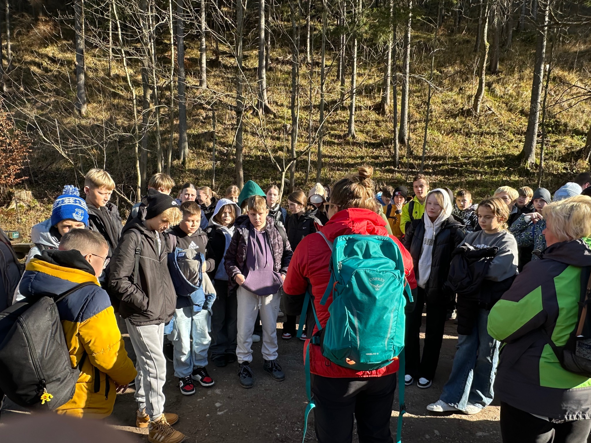 Grupka dzieci i dorosłych słucha przewodnika z plecakiem w lesie podczas słonecznego dnia. Edukacyjna wycieczka na łonie natury.