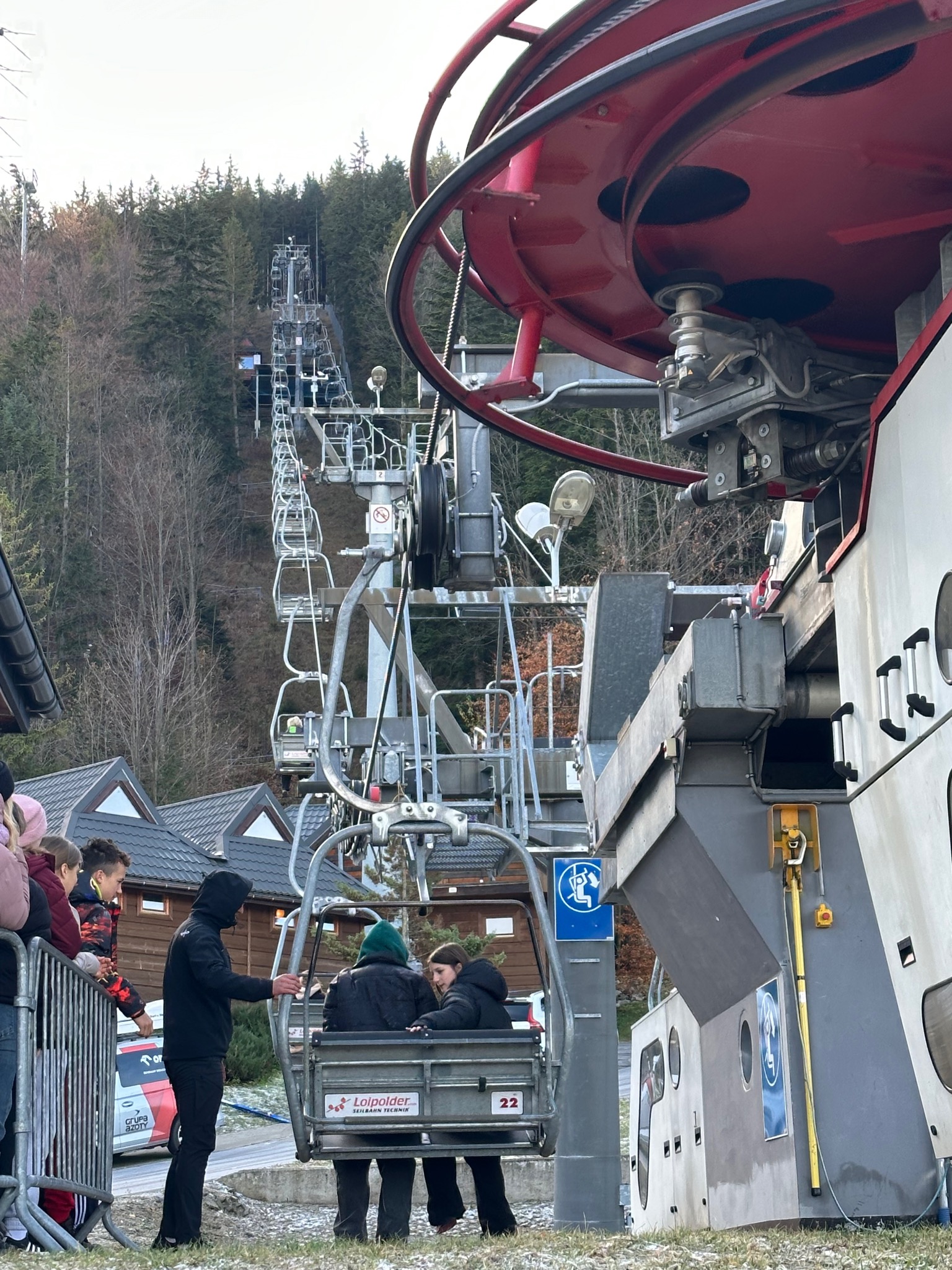 Kolejka krzesełkowa z narciarzami wjeżdżająca na górę, widok z dołu, obok budynek stacji kolejki, w tle las i niebo.