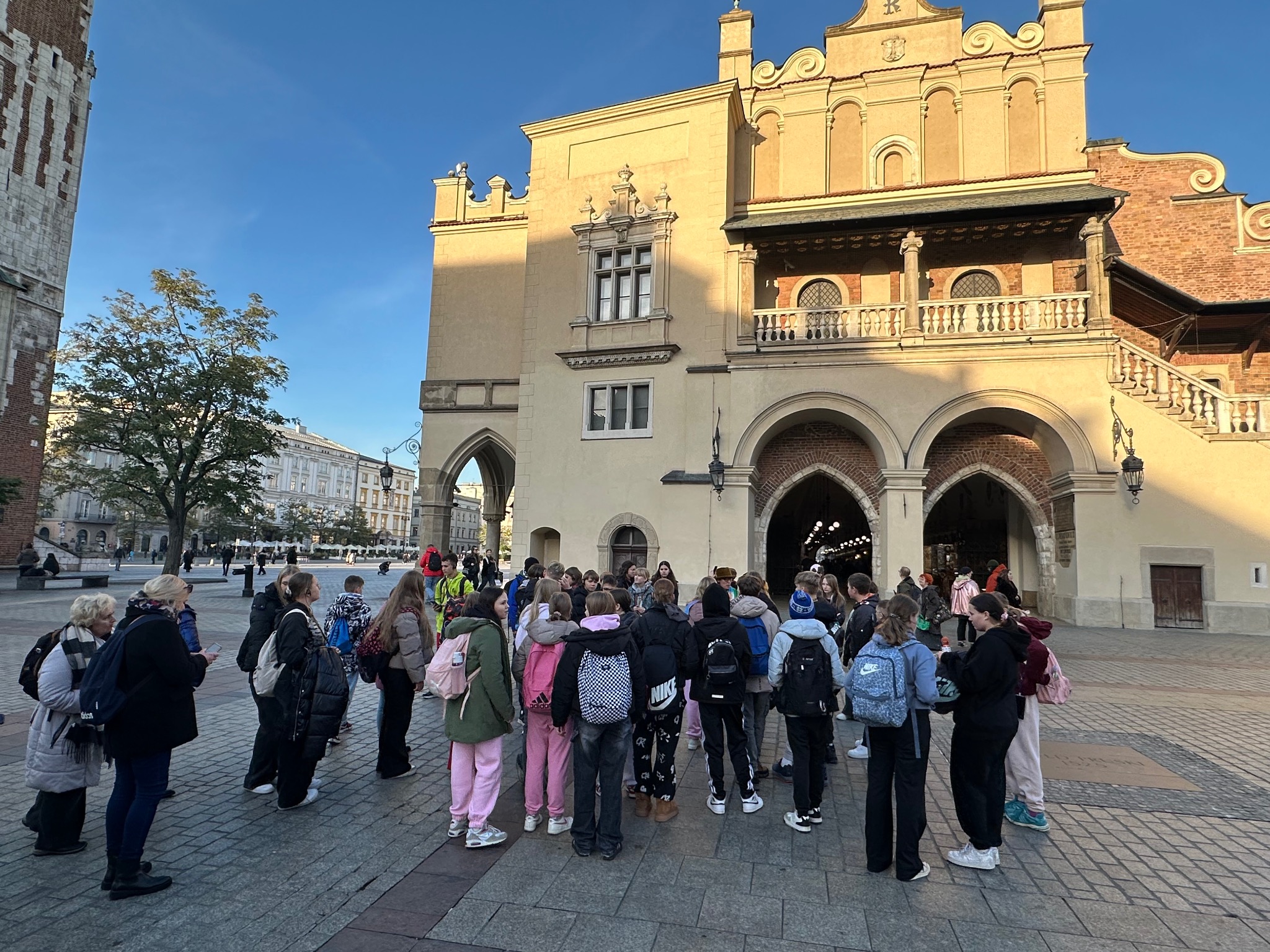 Wycieczka szkolna na Rynku Głównym w Krakowie. Grupa uczniów z plecakami stoi przed Sukiennicami w słoneczny dzień. Widoczna wieża kościoła Mariackiego.