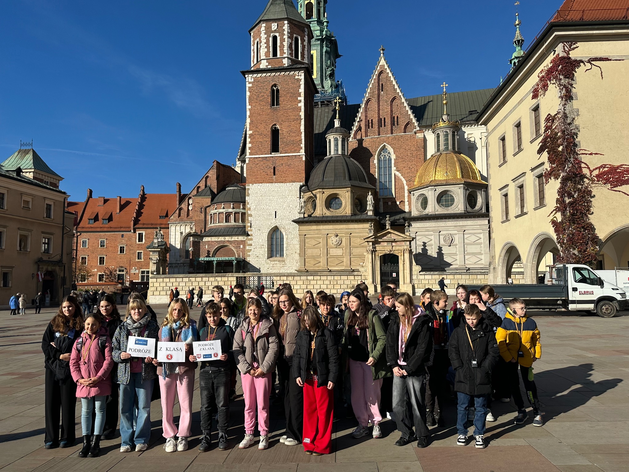 Wycieczka szkolna z Łodzi na tle Wawelu. Grupa uczniów trzymająca tabliczki z napisem 'Podróże z klasą' w słoneczny dzień.