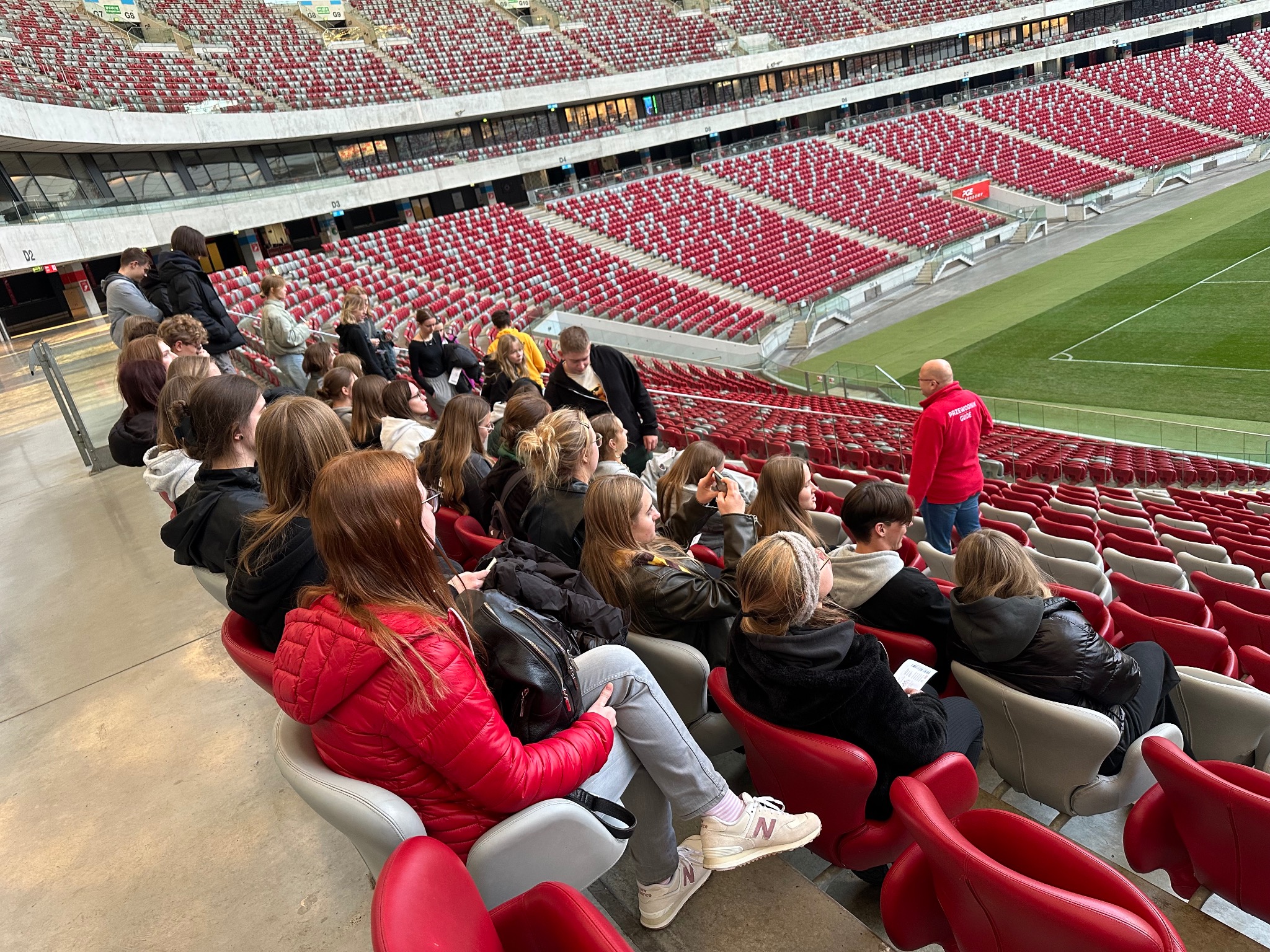 Wycieczka szkolna na stadionie: grupa młodzieży siedzi na trybunach, słuchając przewodnika w czerwonej kurtce. W tle widoczne czerwone krzesełka i boisko.