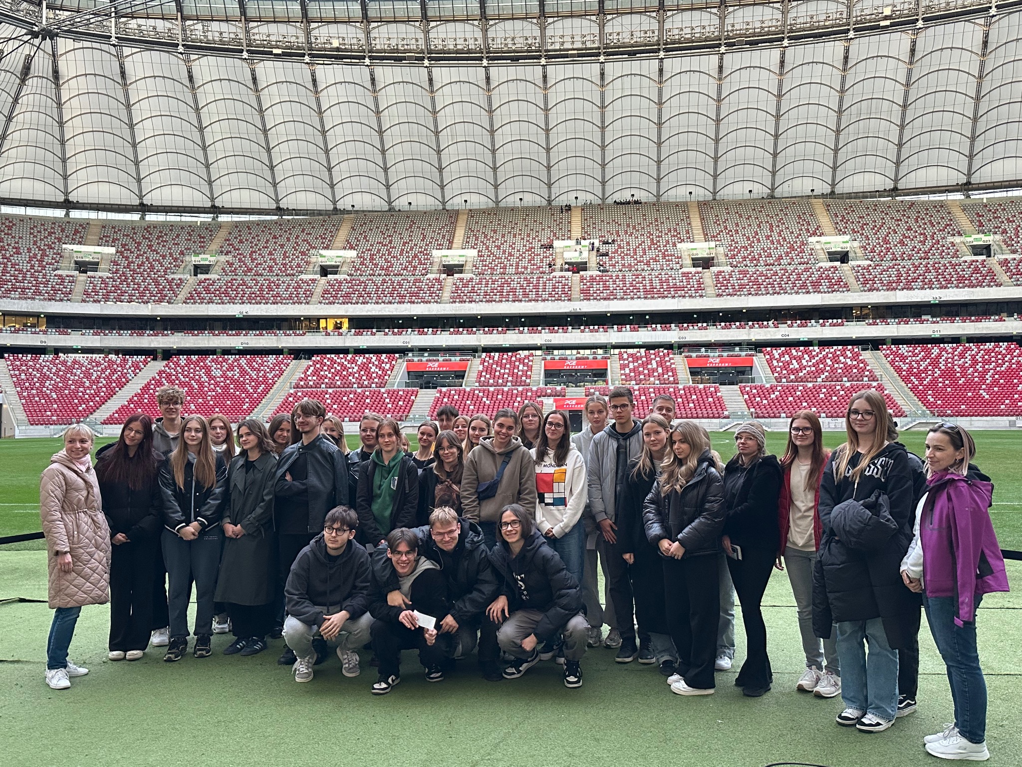Grupa młodzieży na tle trybun stadionu. Wycieczka szkolna z przewodnikiem, zwiedzanie obiektu sportowego. Uczestnicy ubrani w kurtki i bluzy, w tle puste krzesełka.