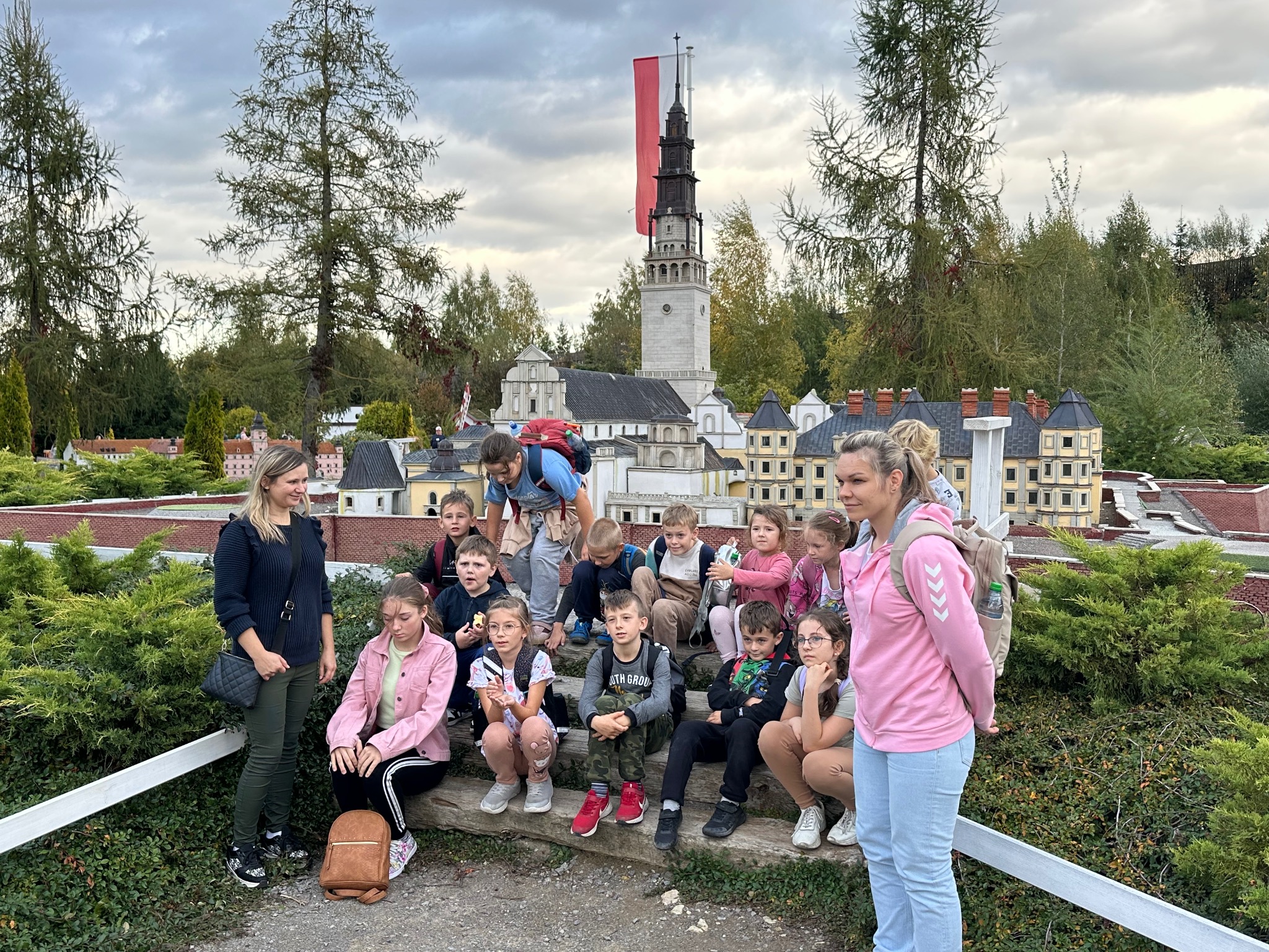 Grupa dzieci z opiekunami zwiedza park miniatur z modelami budowli, w tle wieża z polską flagą. Wycieczka edukacyjna na świeżym powietrzu.