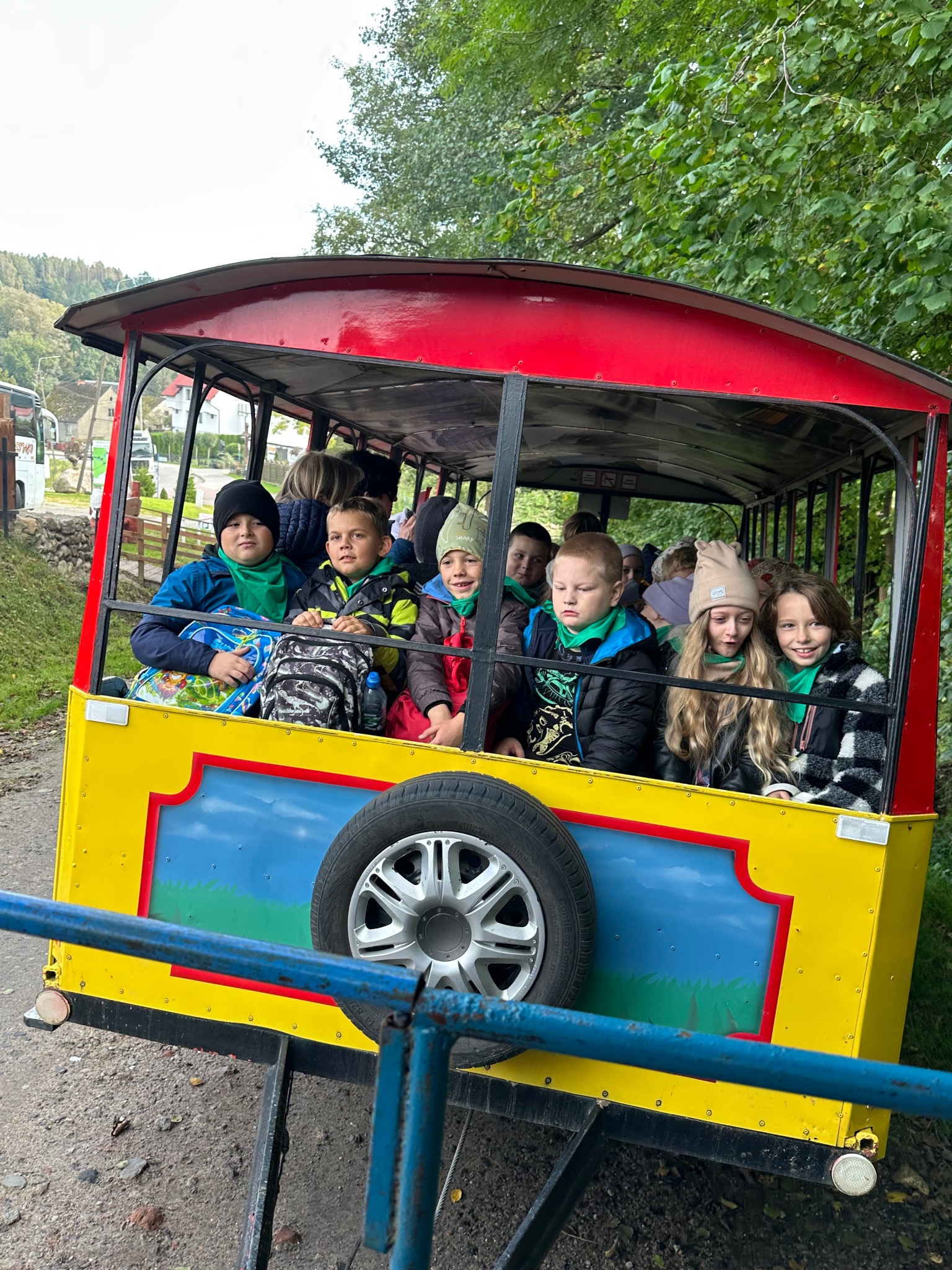 Grupa dzieci w kolorowym wagoniku turystycznym, otoczona zielenią drzew. Wagonik ma żółte boki, czerwony dach i widoczne koło zapasowe. Uczestnicy ubrani w zielone chusty.