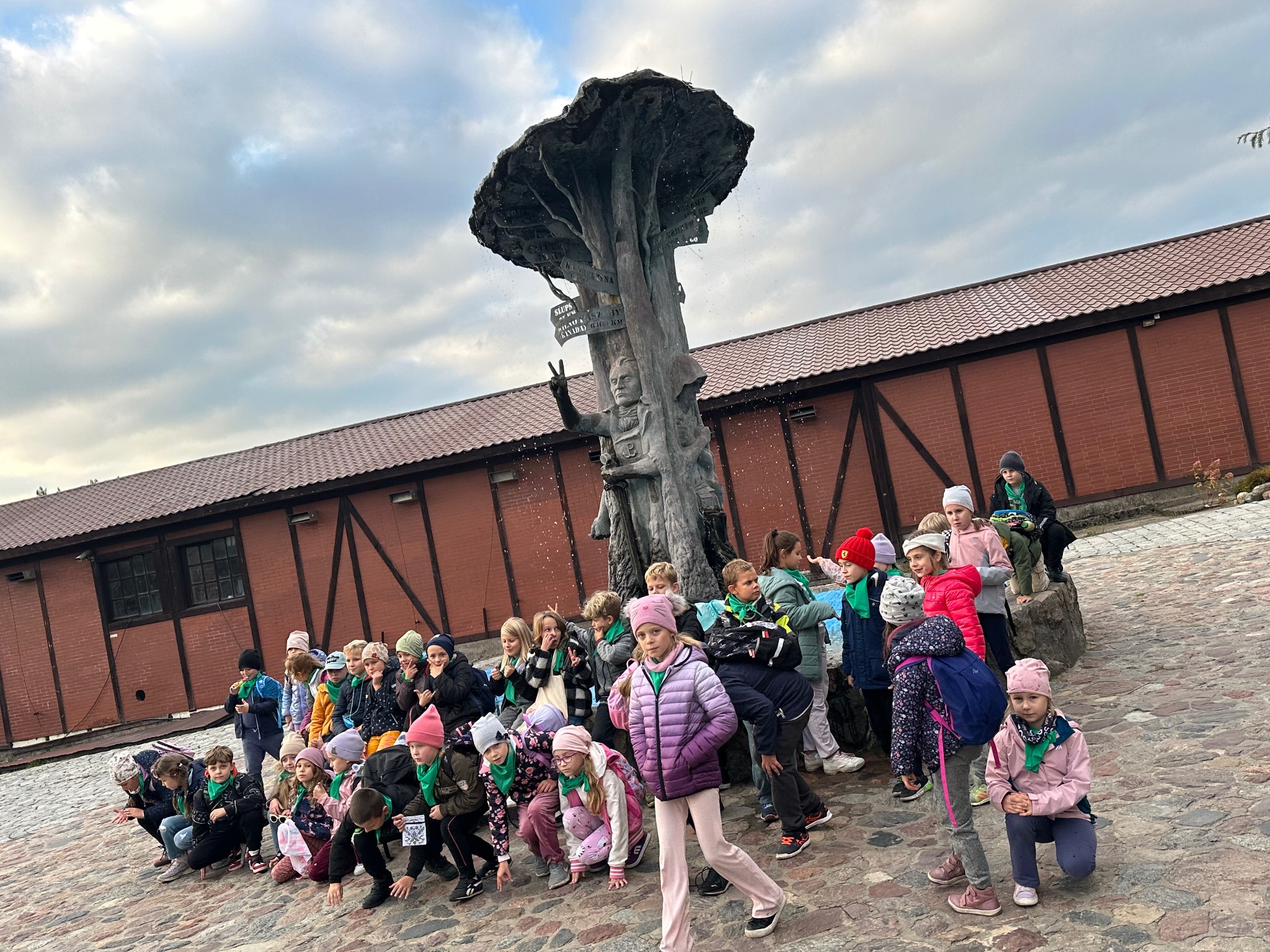 Grupa dzieci w zielonych chustach pozuje przed fontanną w kształcie drzewa z tabliczkami kierunkowymi, ceglany budynek w tle. Pochmurne niebo.