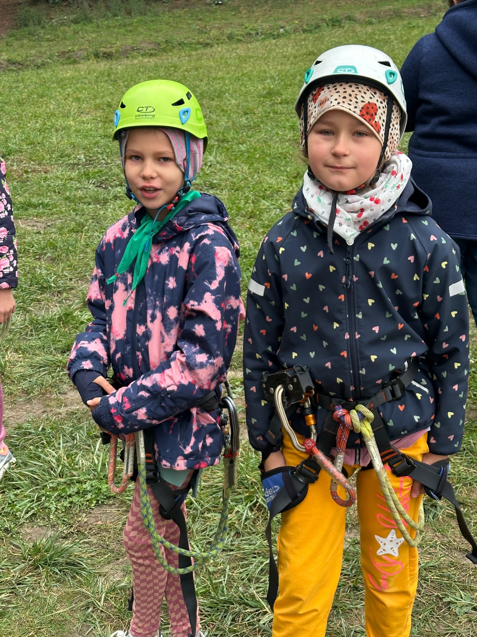 Dwie dziewczynki w kaskach i uprzężach wspinaczkowych stoją na trawie. Jedna ma zielony kask, druga biały. Ubrania kolorowe, w tle zielona trawa i fragmenty innych osób.