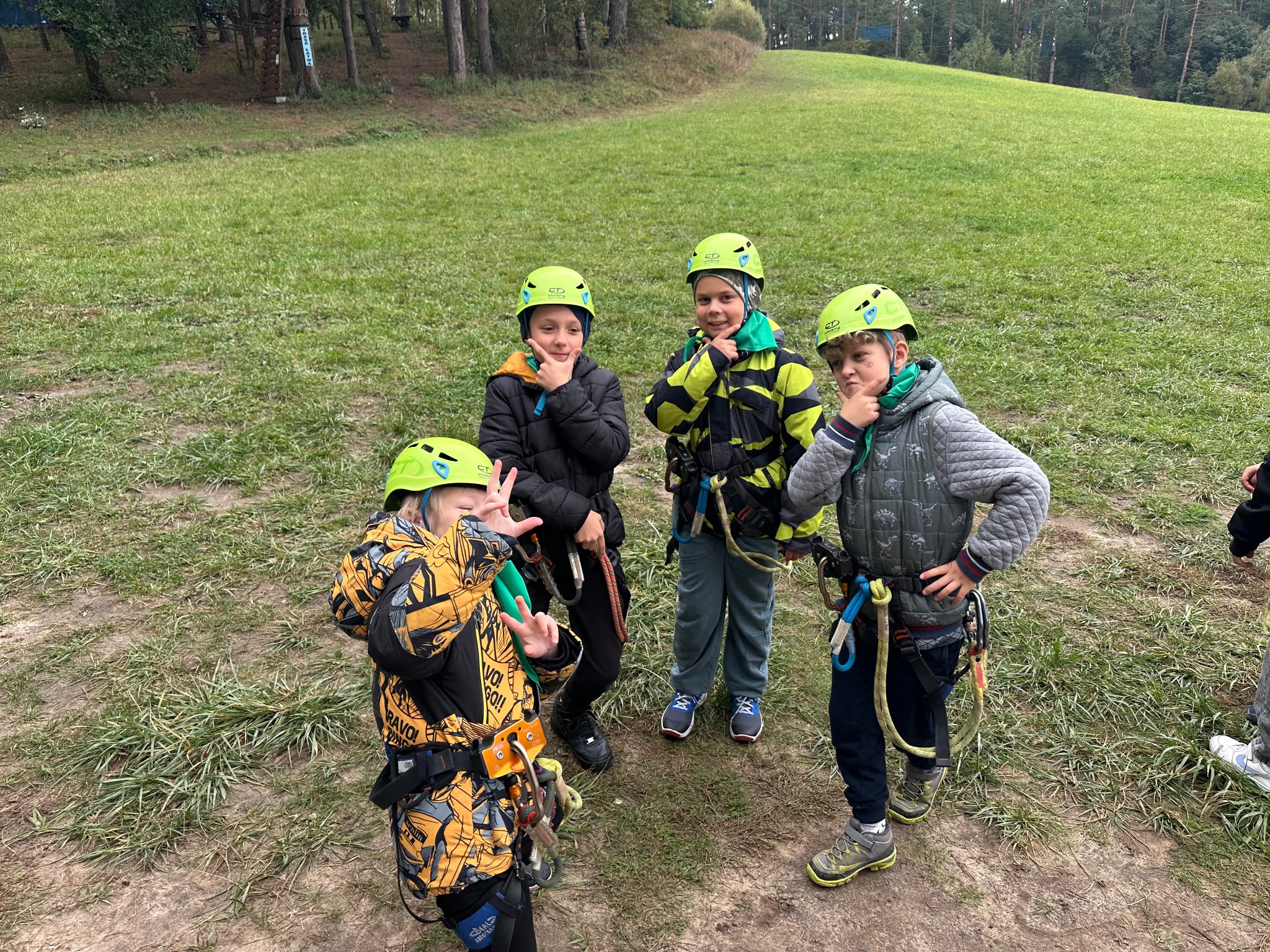 Grupa dzieci w kaskach i uprzężach, przygotowana do aktywności na świeżym powietrzu. W tle widoczny zielony teren leśny, sugerujący park linowy lub obóz.