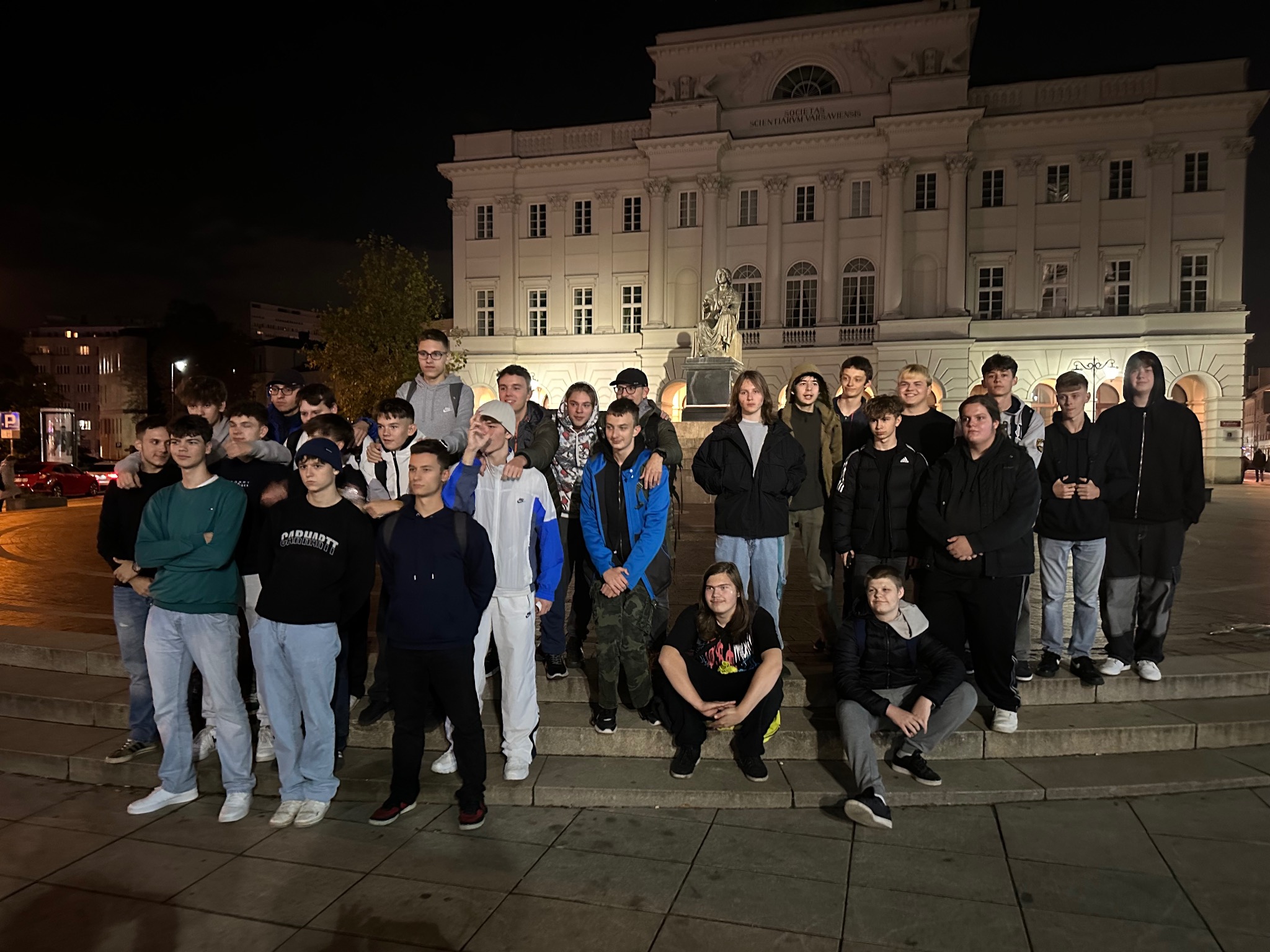 Grupa młodych ludzi pozuje na schodach na tle Pałacu Staszica nocą. Oświetlenie miejskie, młodzież w różnorodnych strojach, widok na architekturę Warszawy.