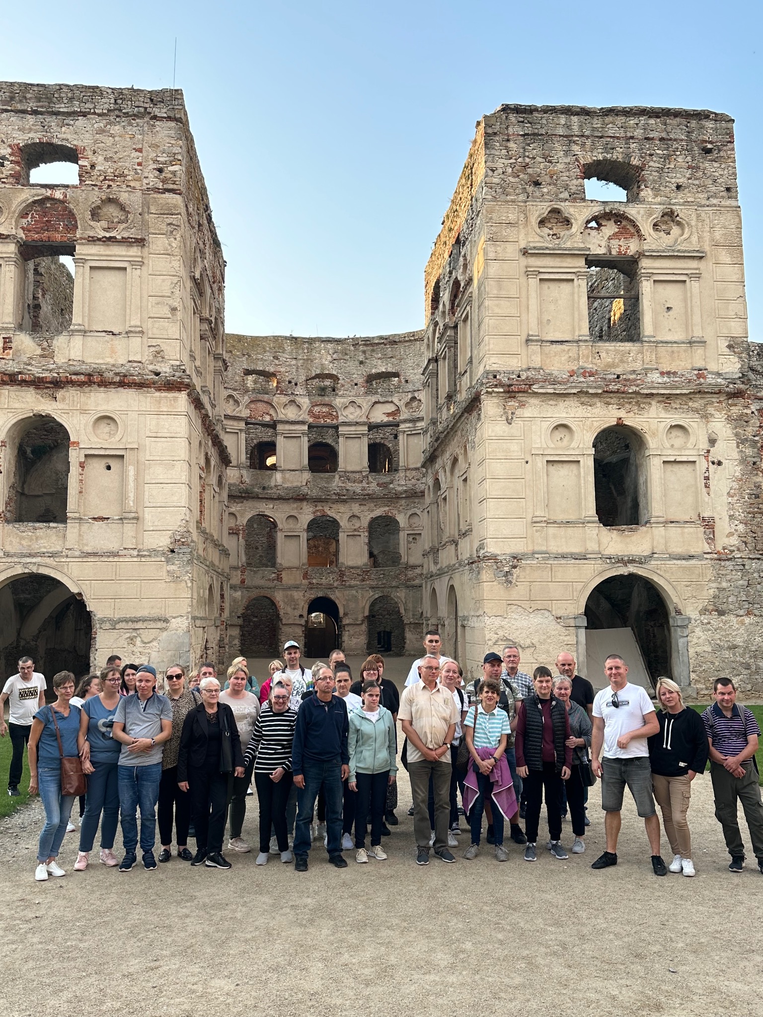 Grupa turystów na tle ruin zamku. Wycieczka zorganizowana, widoczne uśmiechnięte twarze, słoneczny dzień, zabytek architektury.