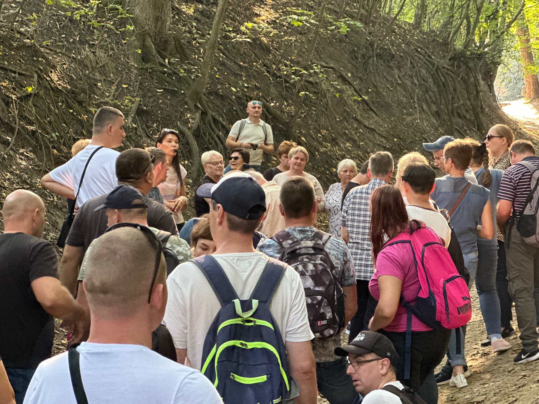 Grupa turystów z plecakami słucha przewodnika w lesie. Ścieżka wznosi się łagodnie, otoczona drzewami i korzeniami. Światło słoneczne przesącza się przez liście.