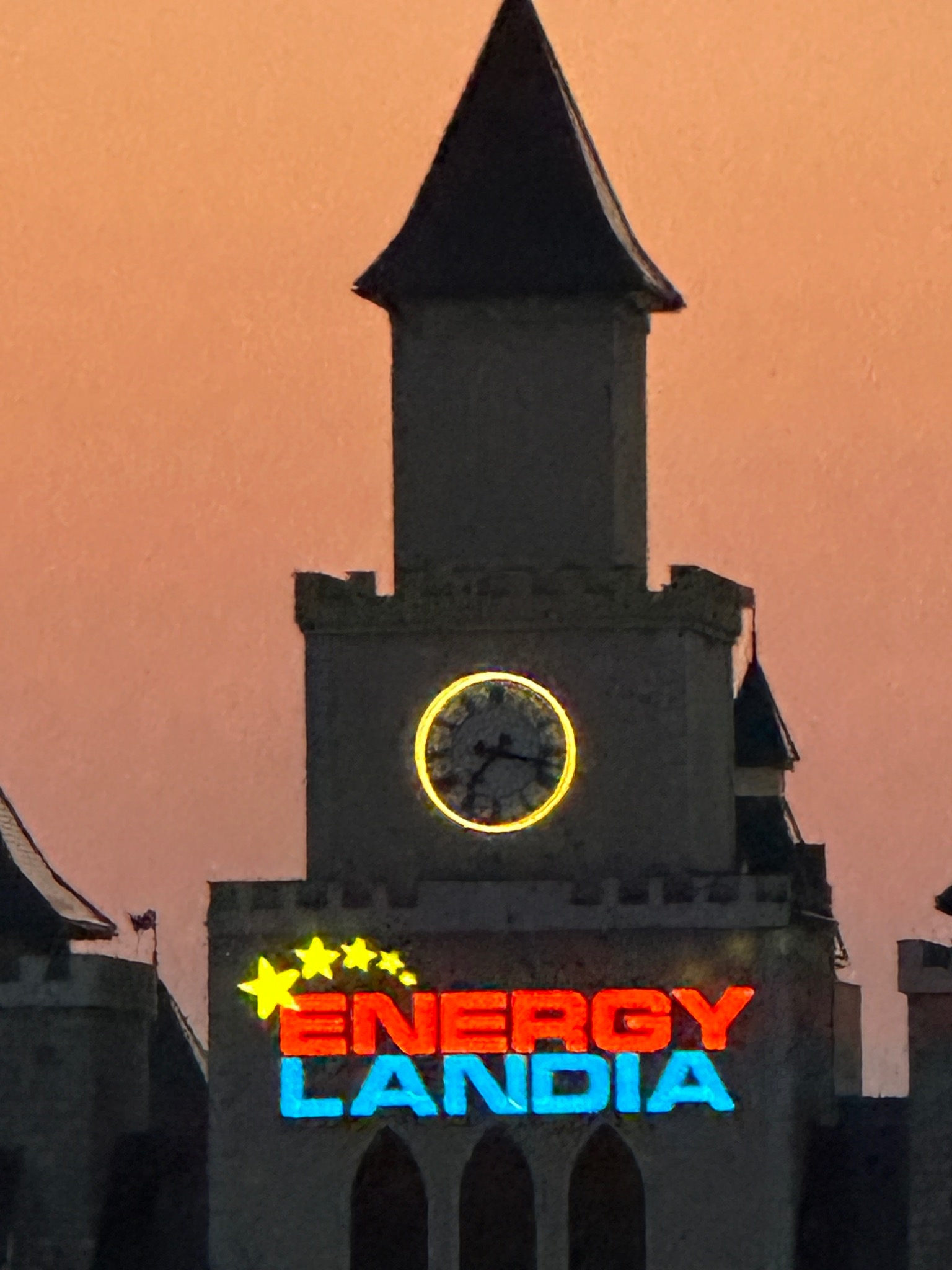 Wieża z zegarem i neonowym napisem Energylandia na tle pomarańczowego nieba o zmierzchu. Widoczne detale architektoniczne zamku.