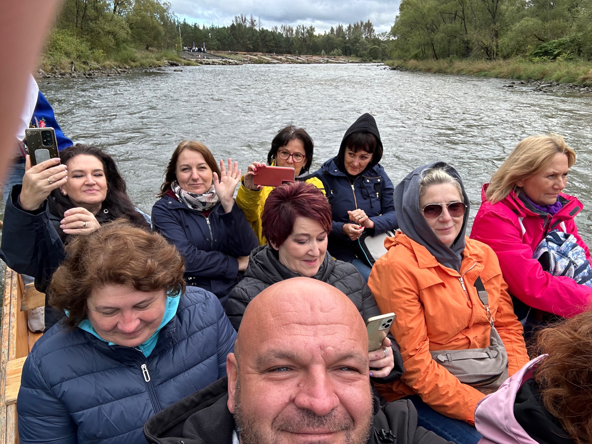 Grupa uśmiechniętych turystów płynie drewnianą tratwą po rzece w pochmurny dzień. Osoby robią zdjęcia telefonami, w tle widoczne drzewa i brzeg rzeki.