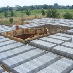 Wieniec stropowy w trakcie budowy, szalunek z drewna otaczający otwór wentylacyjny, widoczne bloczki betonowe i zbrojenie, w tle krajobraz z zielenią.