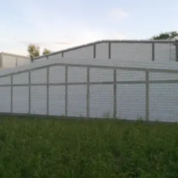 Wznoszenie ścian z białych bloczków na budowie domu, widoczne zbrojenia pionowe i poziome, w tle łąka i drzewa.