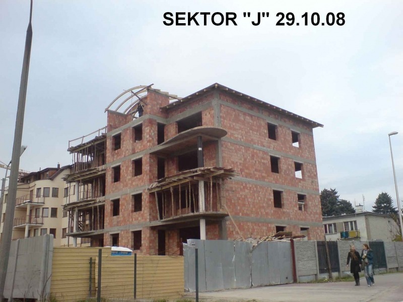 Budynek wielorodzinny w trakcie budowy, widoczne murowane ściany z czerwonej cegły, drewniane szalunki balkonów i elementy konstrukcji dachu, na górze budynku widoczna osoba pracująca, data '29.10.