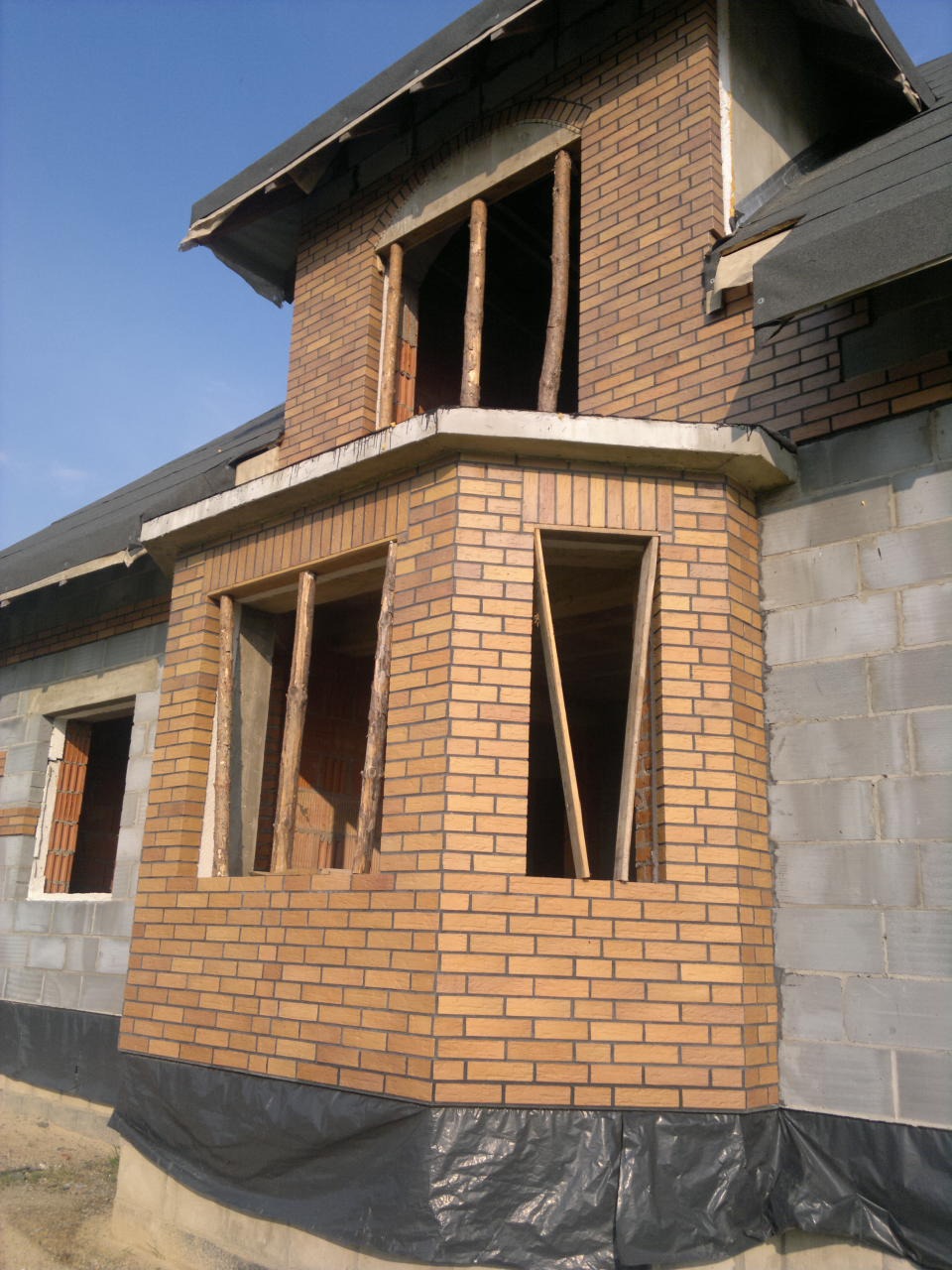 Surowy stan domu w trakcie budowy, z widoczną elewacją z cegły klinkierowej, surową więźbą dachową, oknami bez ram i folią zabezpieczającą fundamenty.