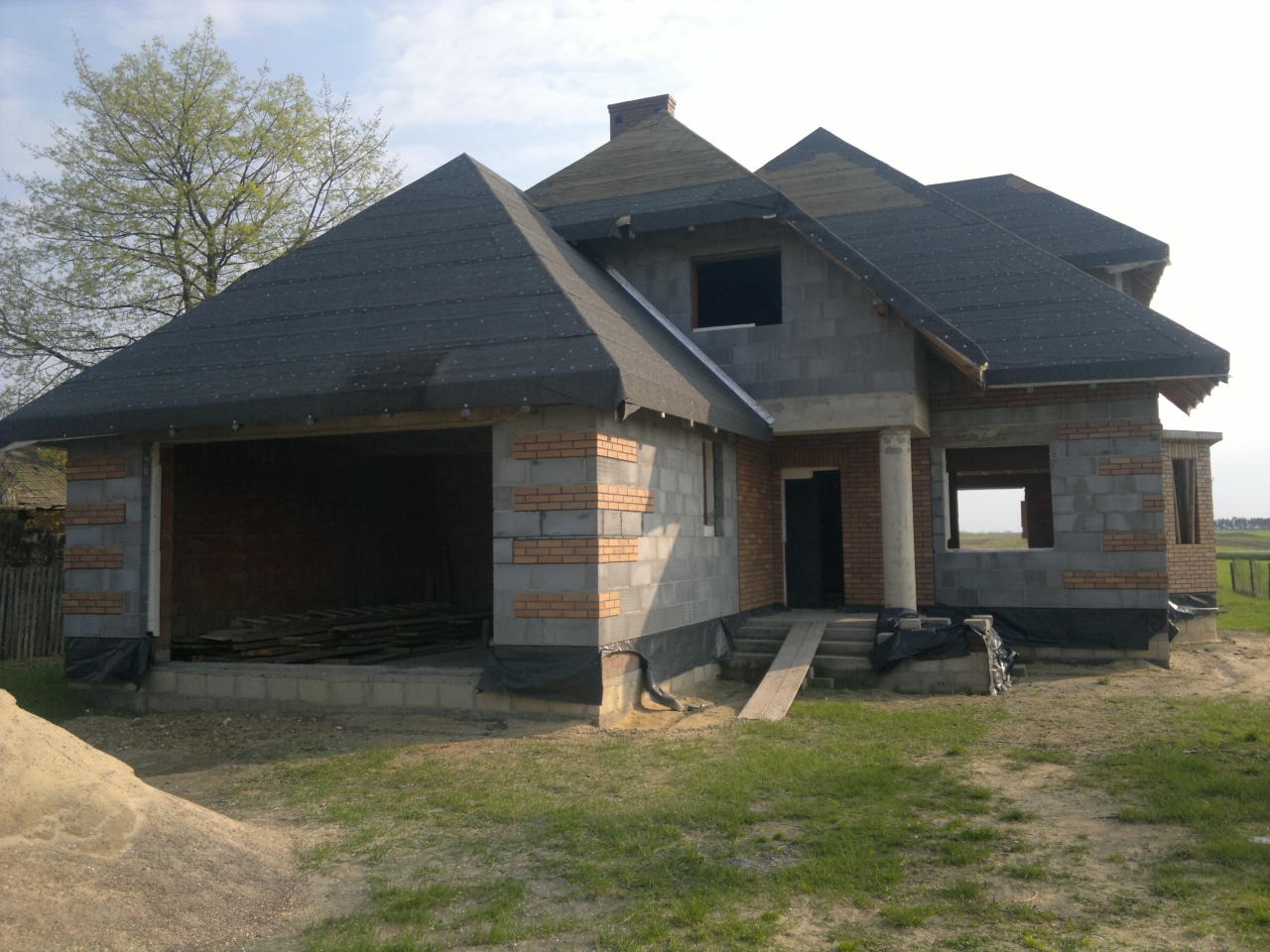 Surowy stan domu jednorodzinnego z garażem w trakcie budowy, ściany z bloczków betonowych i cegły klinkierowej, dach pokryty papą, widoczny stos desek w garażu.