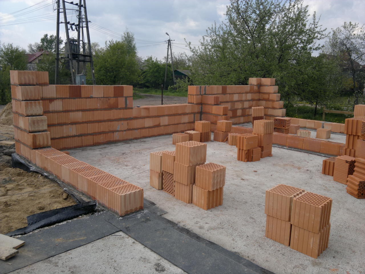 Wznoszenie ścian domu z bloczków ceramicznych, widoczne fragmenty ścian i przygotowane do murowania bloczki na fundamencie z izolacją przeciwwilgociową.