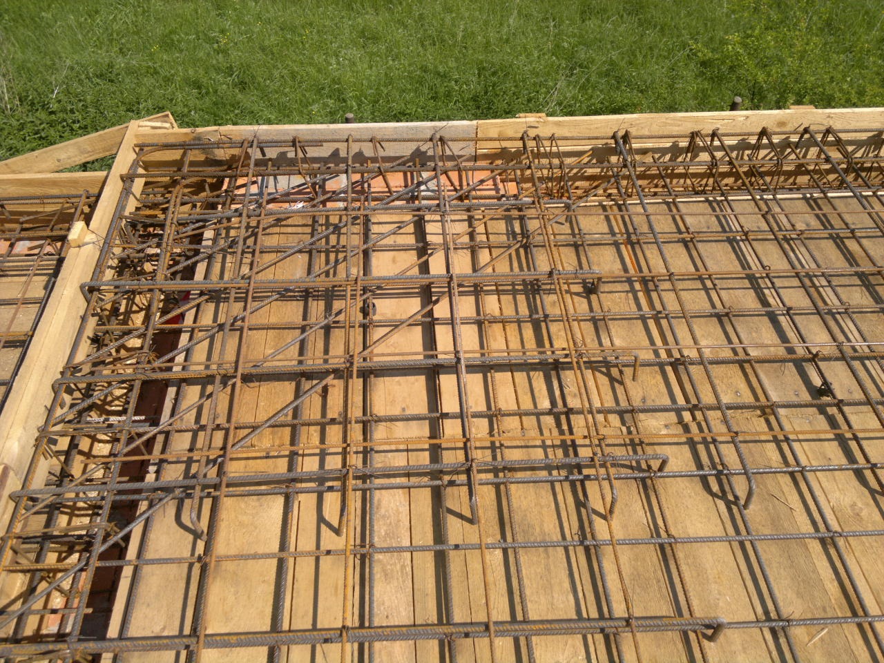 Zbrojenie stropu na deskowaniu z widocznymi elementami drewnianymi szalunku i zielonym tłem trawy.