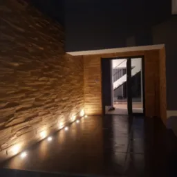 Elewacja zewnętrzna budynku wyłożona kamieniem dekoracyjnym w odcieniach brązu, oświetlona od dołu przez rząd punktowych lamp podłogowych. Widoczne przeszklone drzwi wejściowe z czarną ramą...