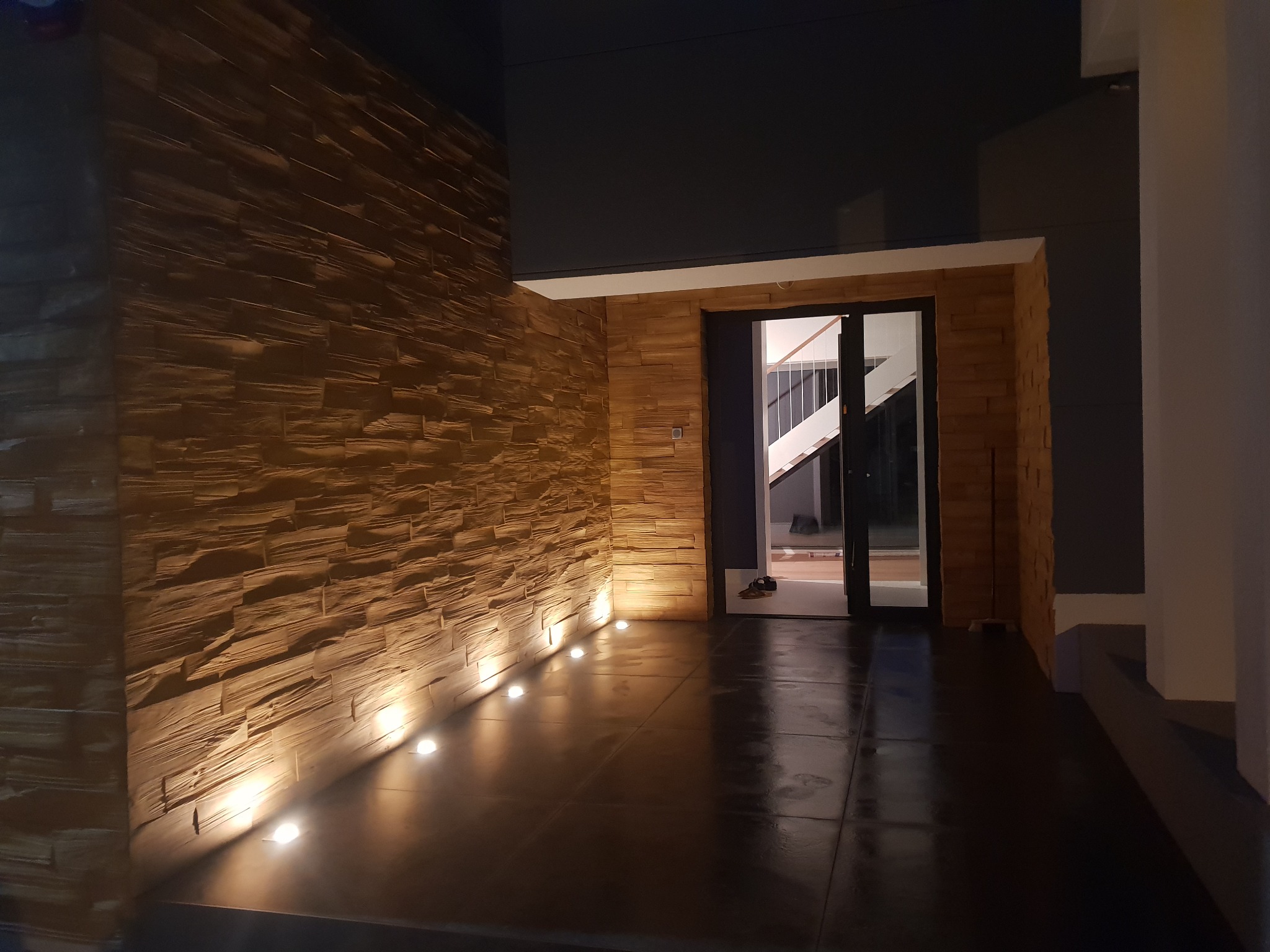 Elewacja zewnętrzna budynku wyłożona kamieniem dekoracyjnym w odcieniach brązu, oświetlona od dołu przez rząd punktowych lamp podłogowych. Widoczne przeszklone drzwi wejściowe z czarną ramą...