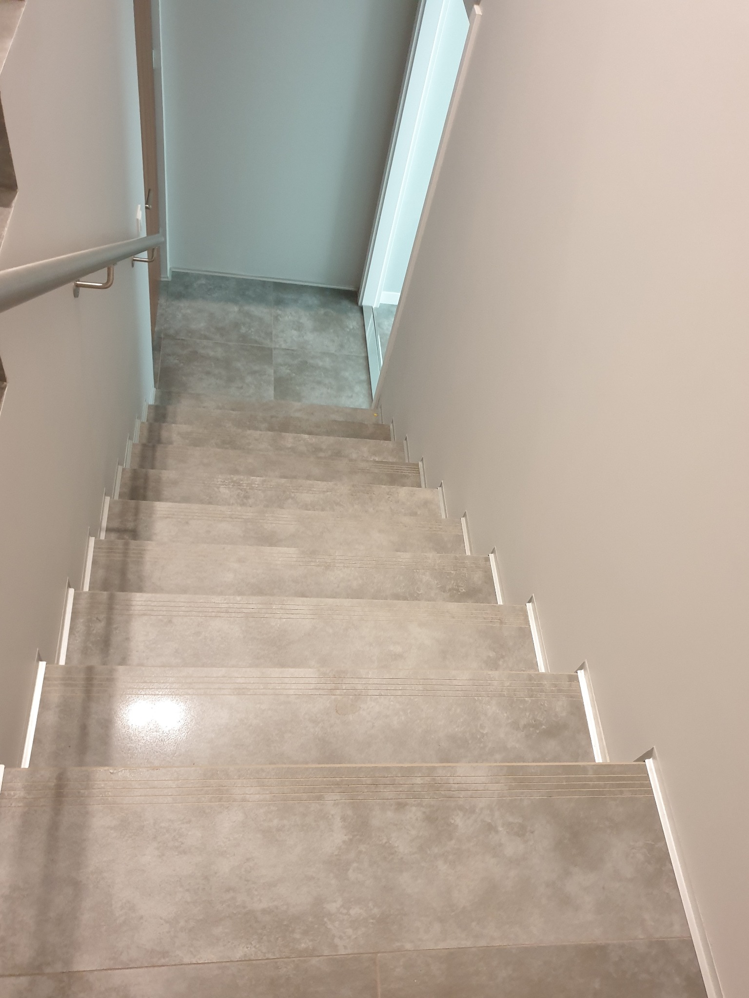 Klatka schodowa wyłożona szarymi płytkami ceramicznymi z metalową poręczą, widok z góry, jasne ściany.