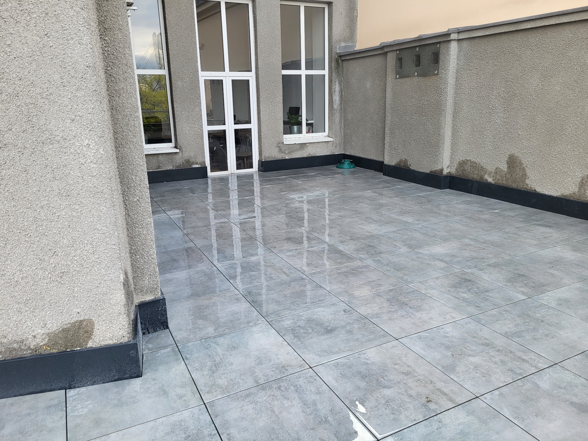 Szare płytki ceramiczne ułożone na balkonie, widoczne okna i ściana z szorstkim tynkiem w tle.