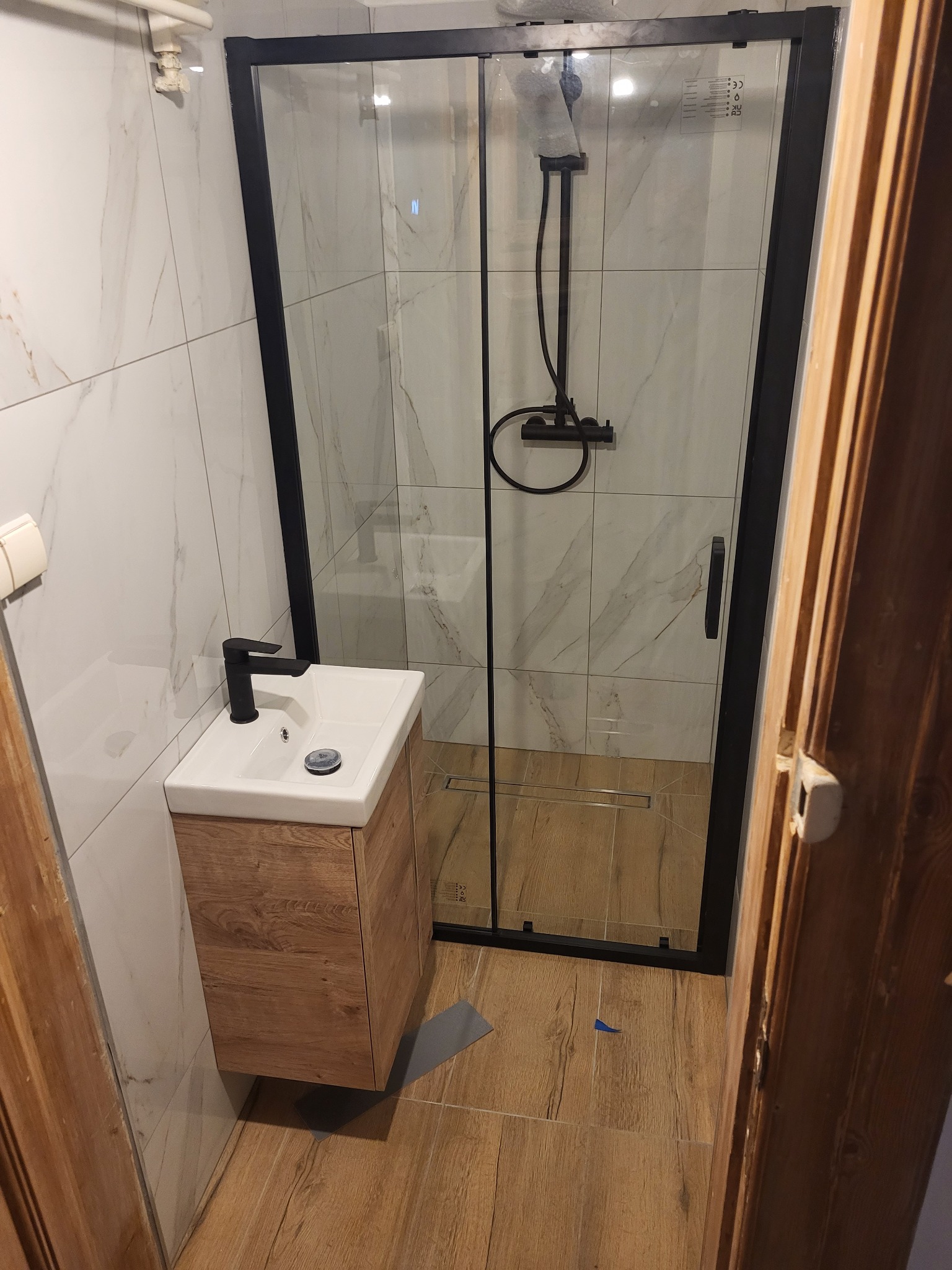 Nowoczesna łazienka w Sulejówku po remoncie: kabina prysznicowa z czarną ramą, umywalka z szafką o drewnianym wykończeniu, marmurowe płytki na ścianach, drewnopodobna podłoga, czarna armatura.