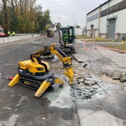 Brokpiec s.c - Żółty robot Brokk z młotem pneumatycznym kruszy asfalt na drodze, w tle operator koparki Bobcat i pracownicy w odblaskowych ubraniach.