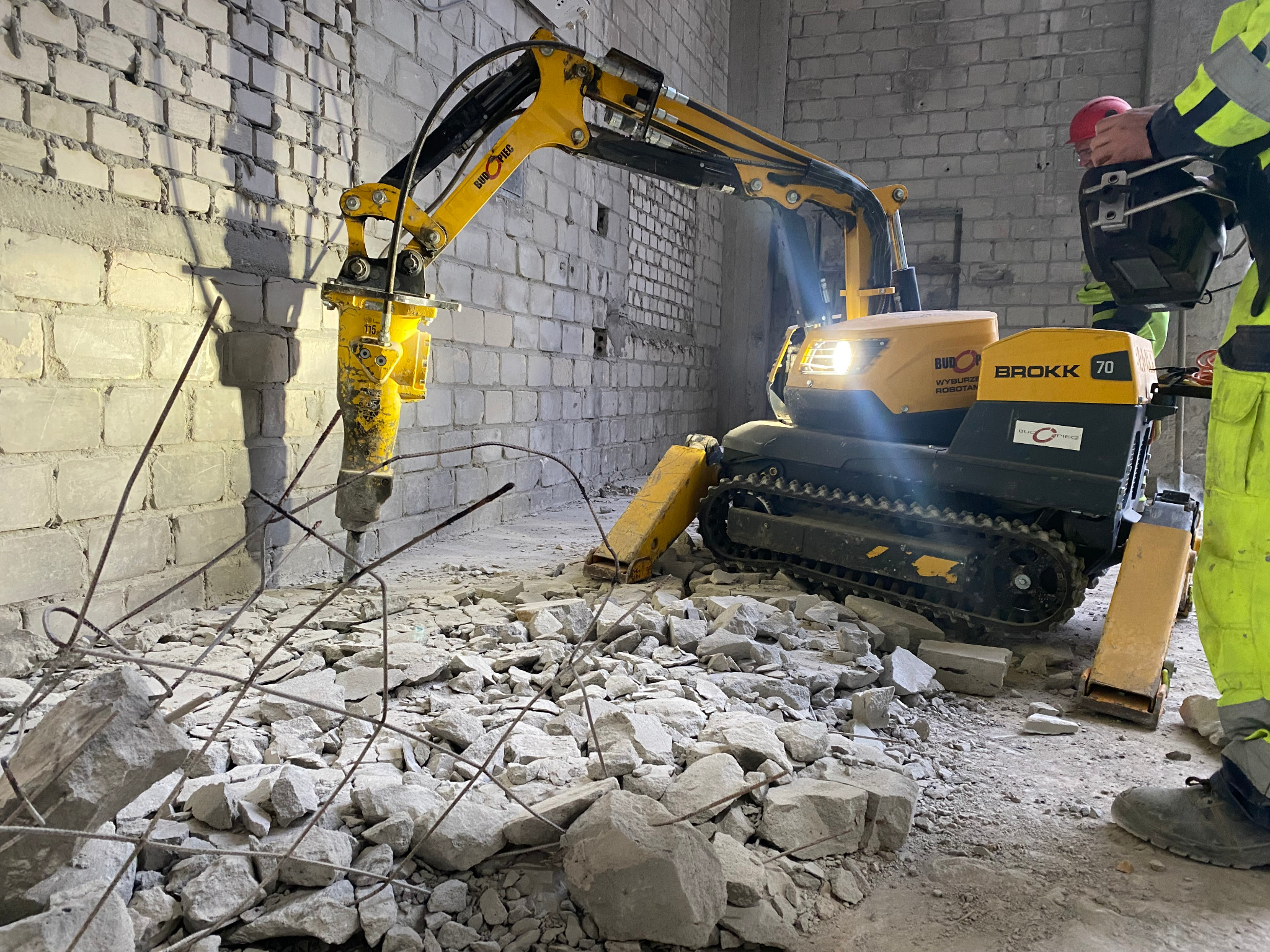 Żółty robot wyburzeniowy Brokk 70 kruszy ścianę z cegły, widoczne gruz i pręty zbrojeniowe, pracownik w kasku i odzieży ochronnej w tle.