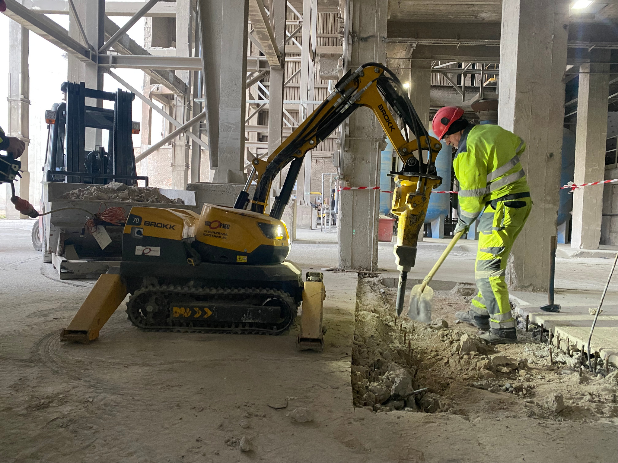 Pracownik w odblaskowej odzieży obsługuje minikoparkę Brokk z młotem hydraulicznym, rozbijając betonową posadzkę wewnątrz hali przemysłowej, w tle wózek widłowy z gruzem.