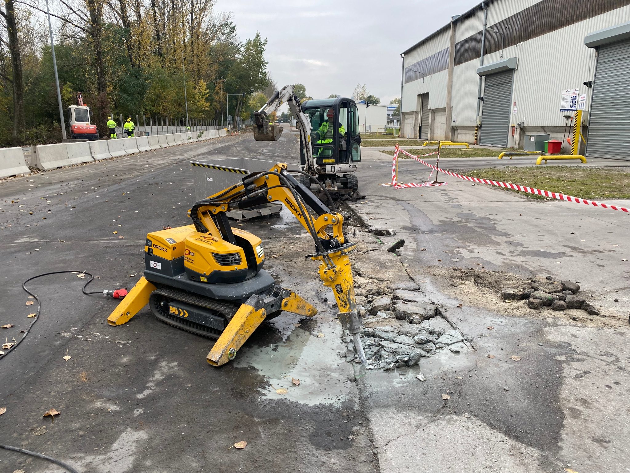 Żółty robot Brokk z młotem pneumatycznym kruszy asfalt na drodze, w tle operator koparki Bobcat i pracownicy w odblaskowych ubraniach.