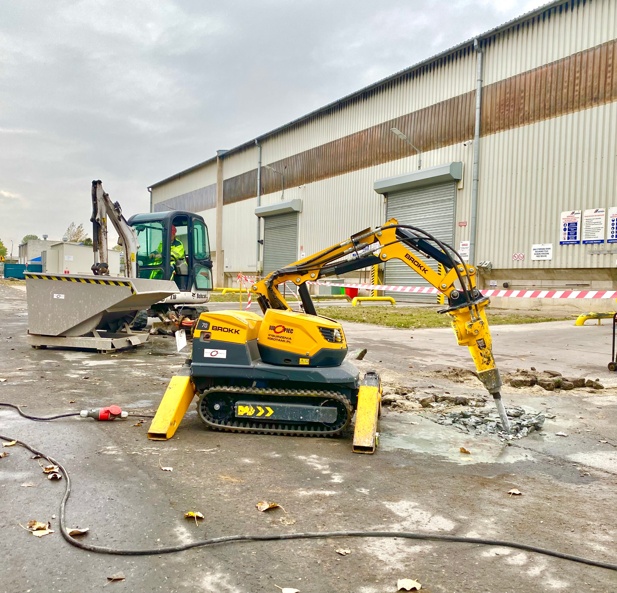 Żółty robot wyburzeniowy Brokk z hydraulicznym młotem rozbija betonową nawierzchnię, w tle budynek przemysłowy i minikoparka Bobcat z operatorem.