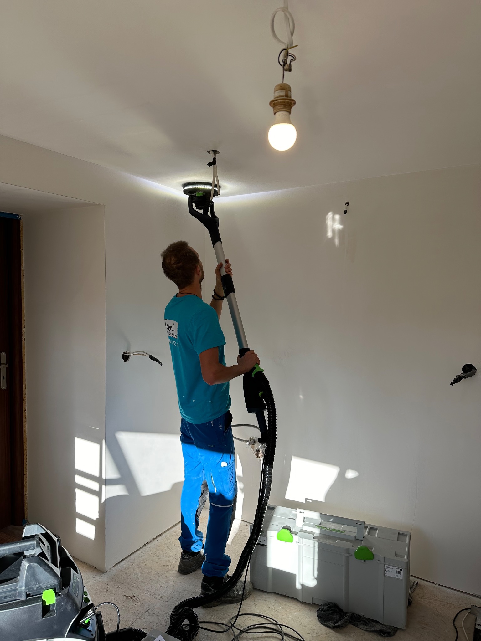 Pracownik szlifuje sufit za pomocą szlifierki z odkurzaczem, widoczne skrzynki narzędziowe Festool, goła żarówka zwisa z sufitu.