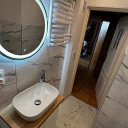 MC Design Jakub Ciuńczyk - Nowoczesna łazienka z marmurowymi płytkami, okrągłym lustrem z podświetleniem LED, białą umywalką nablatową na drewnianym blacie i grzejnikiem drabinkowym.