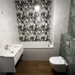 MC Design Jakub Ciuńczyk - Nowoczesna łazienka z drewnianą podłogą, białą armaturą i ścianą w płytkach z motywem roślinnym, widoczne wiszące przewody elektryczne.