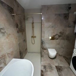 MC Design Jakub Ciuńczyk - Nowoczesna łazienka z marmurowymi płytkami, złotą armaturą prysznicową, podwieszaną toaletą i białą wanną.