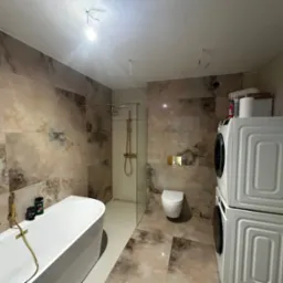 Łazienka w trakcie remontu z marmurkowymi płytkami, białą wanną z złotą armaturą, kabiną prysznicową, toaletą oraz pralką i suszarką w zabudowie, widoczne niedokończone instalacje elektryczne.