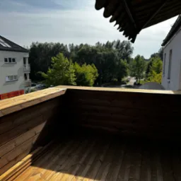 Drewniany taras z widokiem na zieleń miejską, cień markizy na deski, w tle budynek mieszkalny.