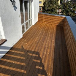 MC Design Jakub Ciuńczyk - Nowo wybudowany taras z desek na dachu budynku, z widocznym drewnianym ogrodzeniem i białymi drzwiami balkonowymi, oświetlony promieniami słonecznymi rzucającymi cienie na deski.