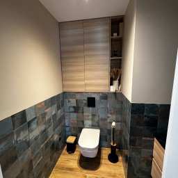 MC Design Jakub Ciuńczyk - Nowoczesna toaleta z płytkami imitującymi rdzę, drewnianą podłogą i szafkami, uzupełniona o czarne akcenty i dekoracje.